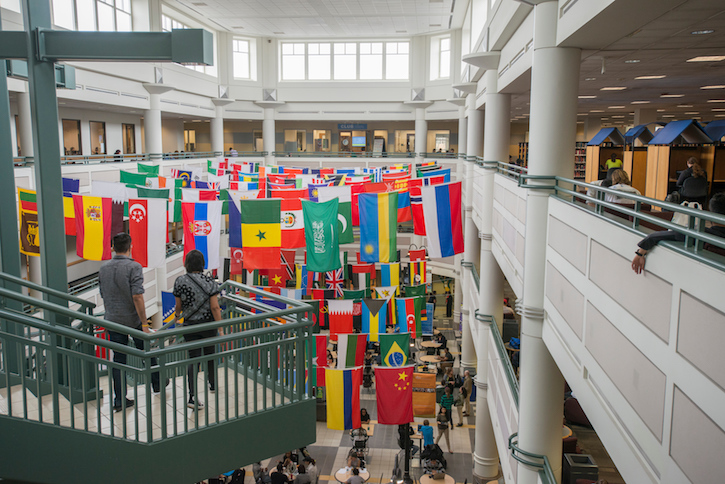 Photo of a bustling university student center. On the right of the frame, two individuals walking down a green staircase on the right of the frame. On the left of the frame is a white balcony wall with individuals sitting at tables alongside it. In the middle of the frame, rows upon rows of flags from across the world hang over an indoor courtyard.